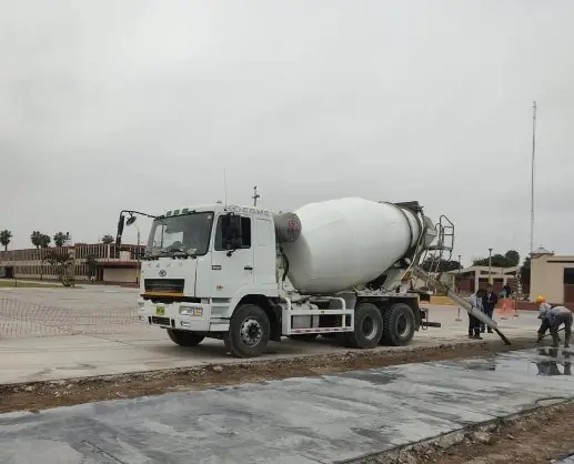 Mixer blanco descargando concreto por su canaleta en una losa a nivel del suelo, con obreros y una explanada de concreto al fondo
