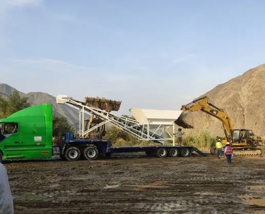 Planta de concreto móvil sobre una cama baja en un terreno de obra, junto a una excavadora